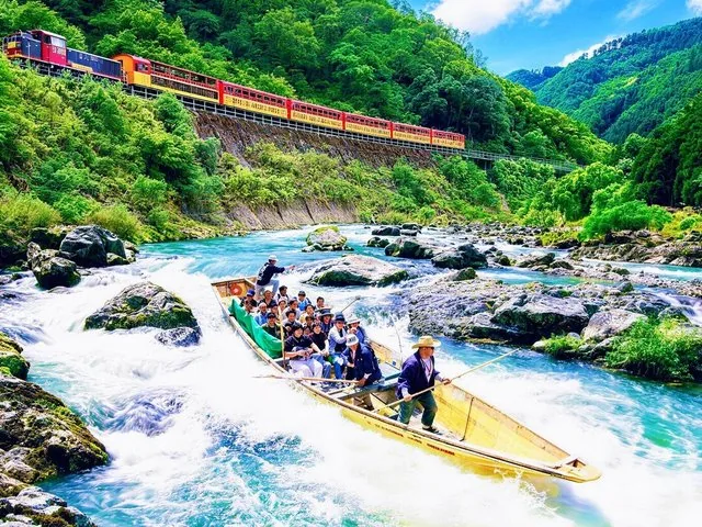 A boat full of passengers travels down the Hozugawa River alongside a train