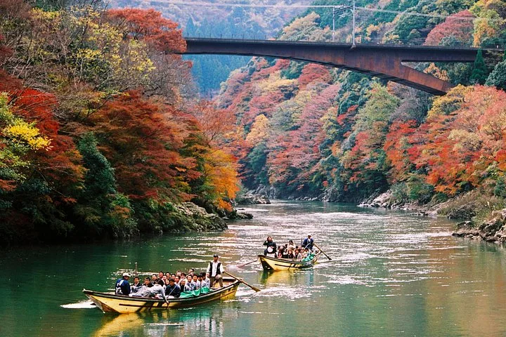 Two boats float down Hozugawa River surrounded by autumn foliage and a bridge