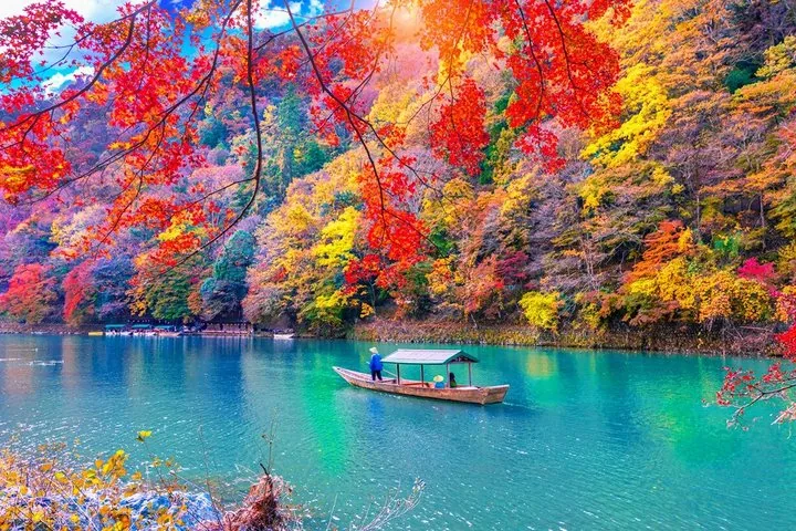 A traditional boat glides on vibrant blue water surrounded by autumn foliage